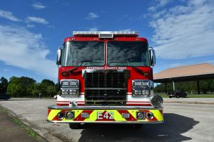 This is an image of the front of Engine 42. The bright red vehicle has a stripe that goes around the front cab, just above the front bumper. The Bumper is striped fire engine red and reflective yellow, with E42 in the center. Chome accents adorn the front around the headlights, emergency lights, mirrors, sirens, and horns. A bright blue sky with a few light wisps of clouds are in place above the vehicle.