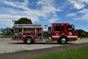 Engine 42, a brand new fire engine is centered in the image. the image shows the passenger (right) side of the vehicle, with all the utility doors open. The emergency extraction equipment is all on display as light, lazy clouds coast through a bright blue sky.