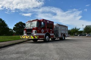 Engine 42 is shown from the front three quarters, drivers side view. The massive bumper sticks out a few feet in front of the vehicle, like a giant chrome ledge - a siren perched on the front left corner. The side of the vehicle has the ESD logo on the driver door, and ENGINE 42 on the back door. The doors to the emergency equipment are closed showing the brand three new aluminum doors, with the white stripe continuing onto the forward door, where it makes a backward Z and then continues across the remaining two roll doors. A round decal about a foot in diameter is placed near the bottom of the last door with 911 clearly visible