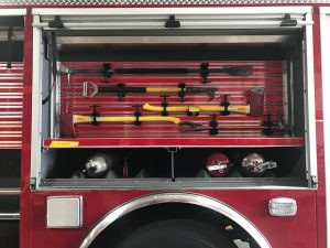 A clear closeup image of one of the emergency extraction tool locations located above the rear passenger wheel shows a number of devices, including three different halebrands and two types of axes on adjustable mounts. A bin just below has seperated dividers to hold up to 8 fire extinguishers. The space is shown with one in the second to last (most left) two fire extinguishers in the second and third slots (fron the right of the photo).