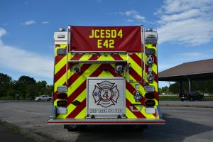 A direct rear view of Engine 42 shows a large lower platform for fire fighters to stand on. It's diamond plate surface glitters in the sun. An aluminum rollup door containing fire hoses is centered just above the platform and has a large decal of the Jefferson County Emergency Services District #4, with the words KEEP BACK 500 FEET right below it. The remainder of the back of the engine alternates with fluorescent yellow and fire engine red chevrons. Assorted backup, brake and emergency lights... and numerous other chrome fixtures adorn the back of the new engine.