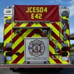 A direct rear view of Engine 42 shows a large lower platform for fire fighters to stand on. It's diamond plate surface glitters in the sun. An aluminum rollup door containing fire hoses is centered just above the platform and has a large decal of the Jefferson County Emergency Services District #4, with the words KEEP BACK 500 FEET right below it. The remainder of the back of the engine alternates with fluorescent yellow and fire engine red chevrons. Assorted backup, brake and emergency lights... and numerous other chrome fixtures adorn the back of the new engine.